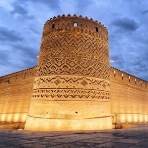 The wall of Karim khan Citadel and many locals and tourists around it, there's 3 flags of Iran in front of the entrance, Iransense Travel Agency